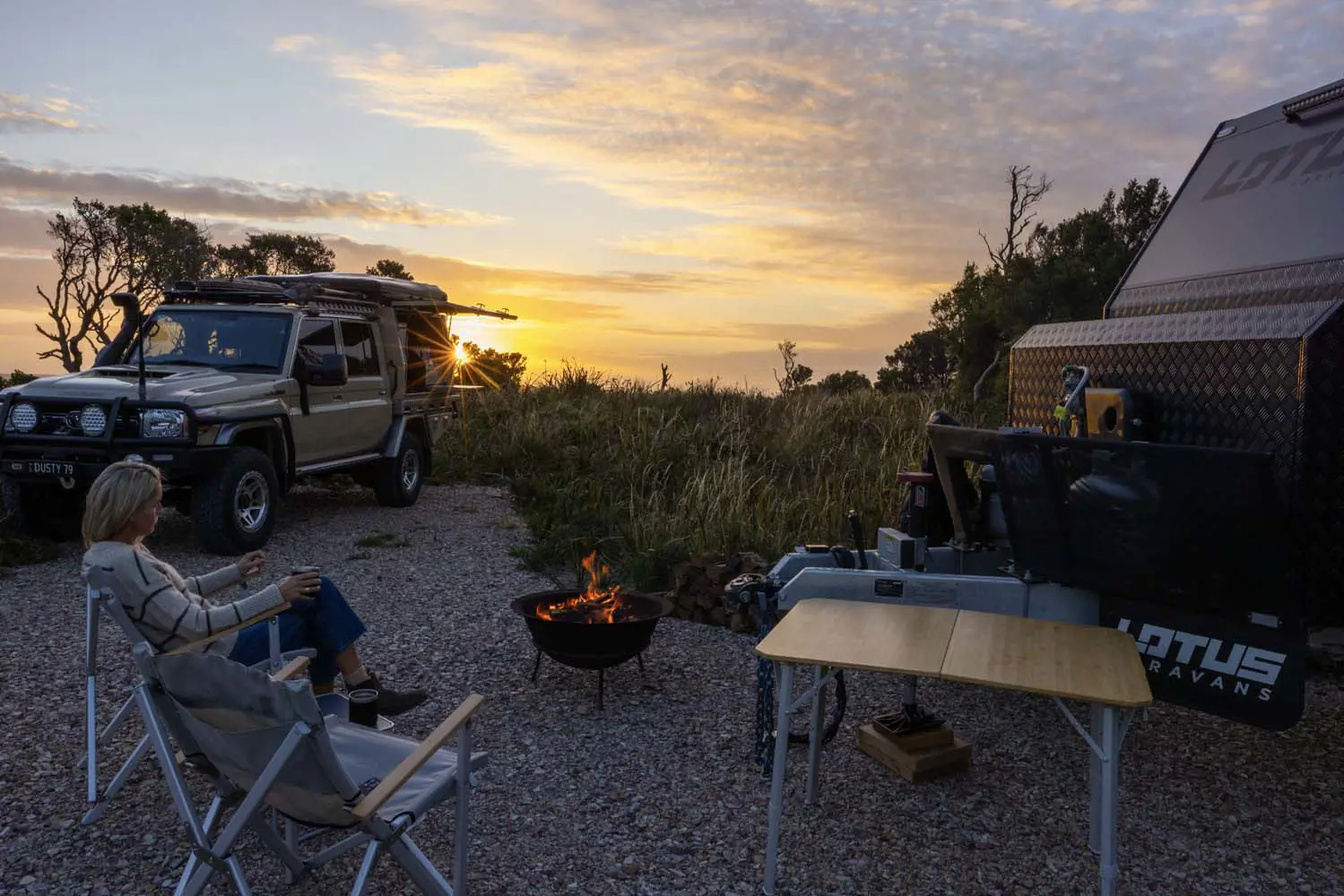 An outdoor campground setup at sunset time, with a firepit and comfy chairs next to a caravan.