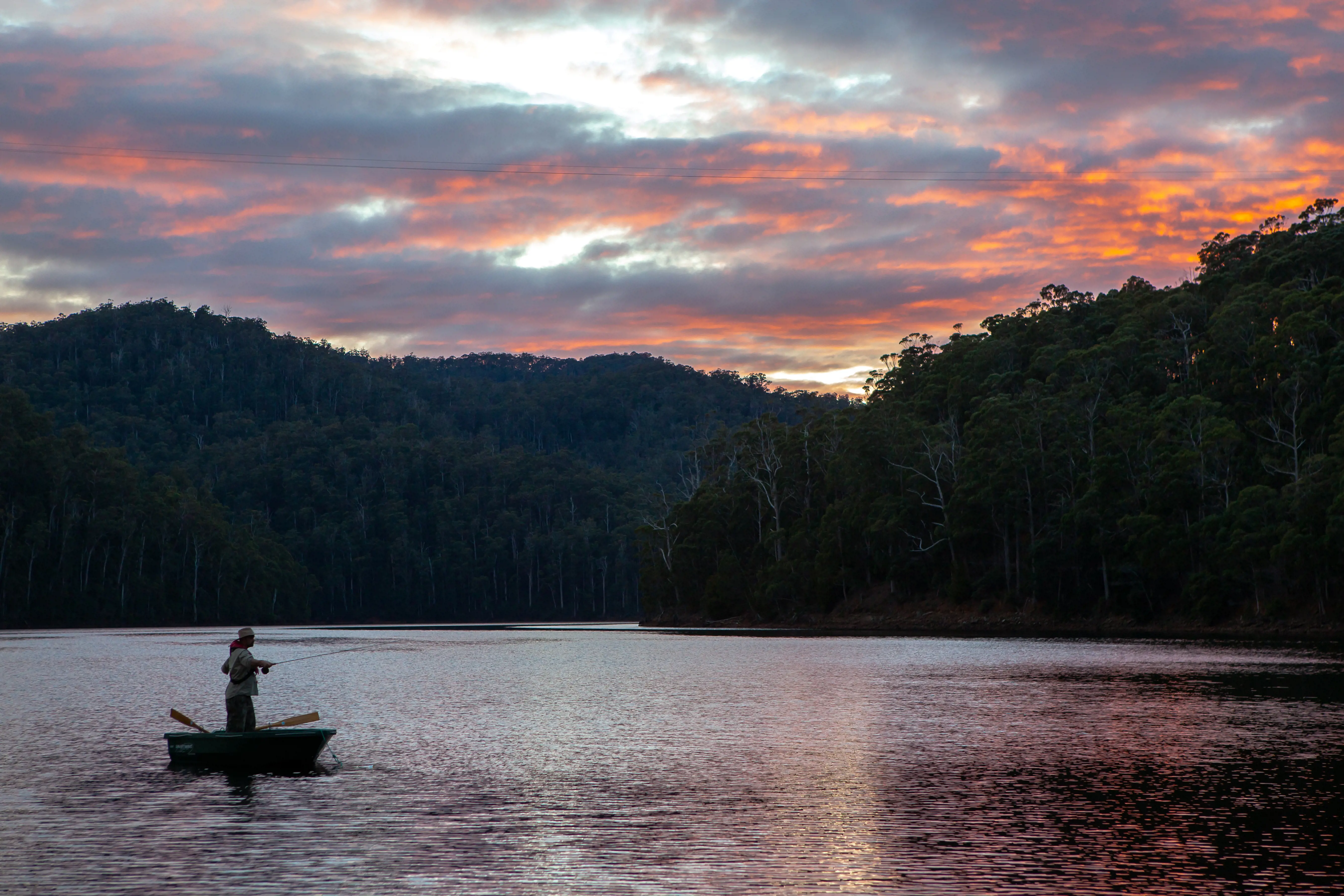 A person stands in a boat with a fishing rod on a wide lake, surrounded by steep bushy banks. The sky and clouds are golden pink at sunset.