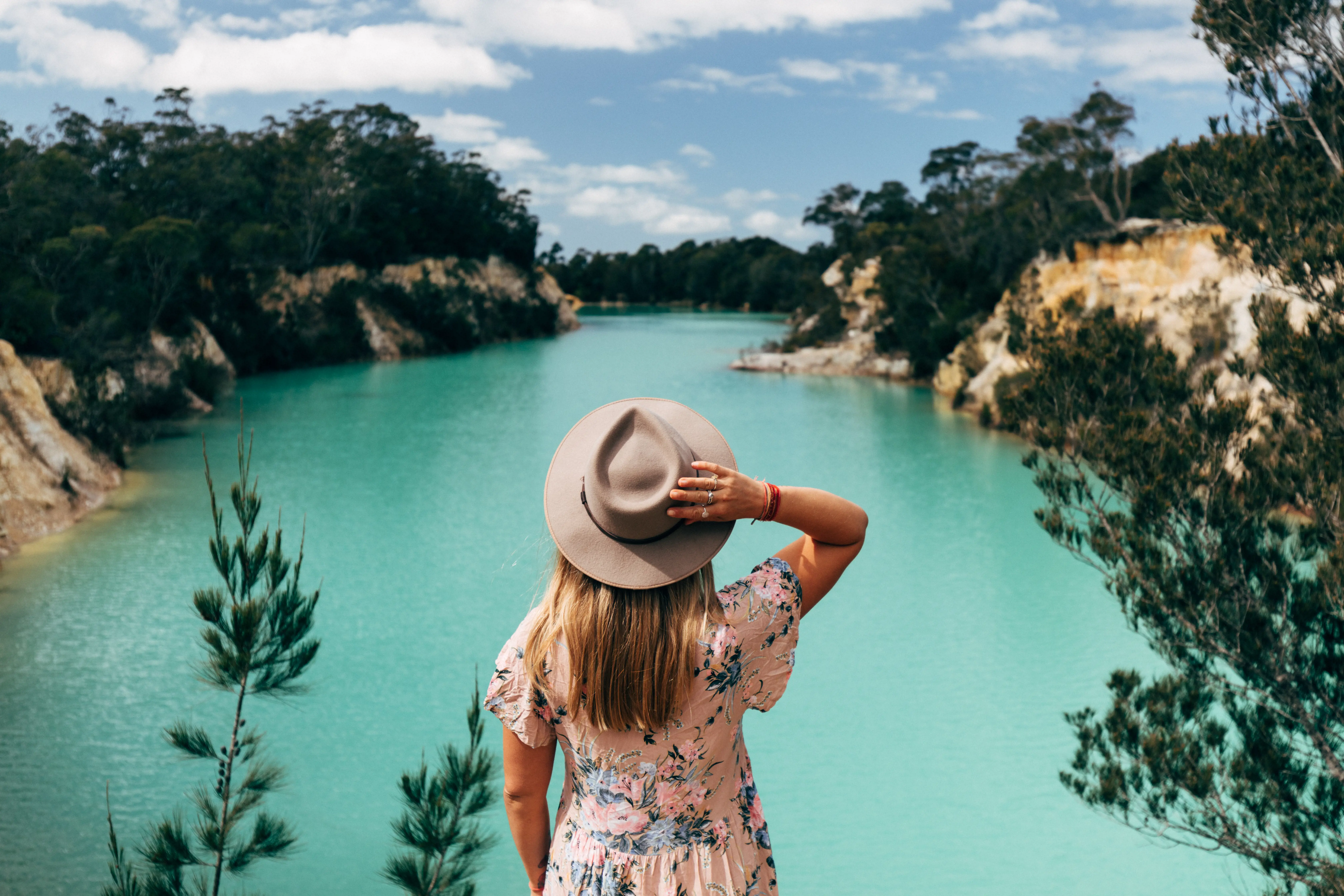 A woman holding her hat onto her head stands in front of a vivid aqua blue lake.