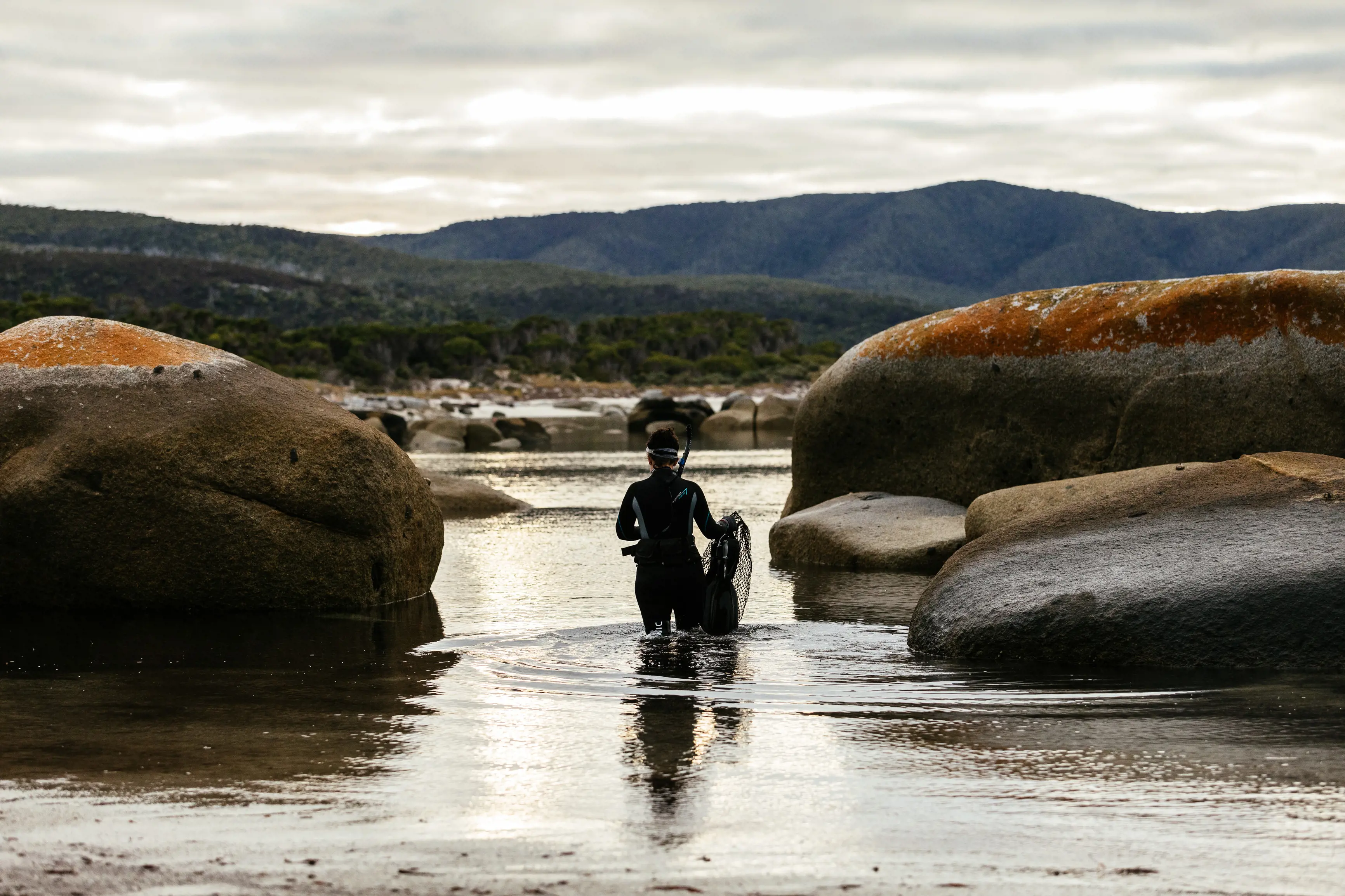 A person in a full wetsuit and snorkel holds a net standing thigh-deep in ocean water between some large rocks.