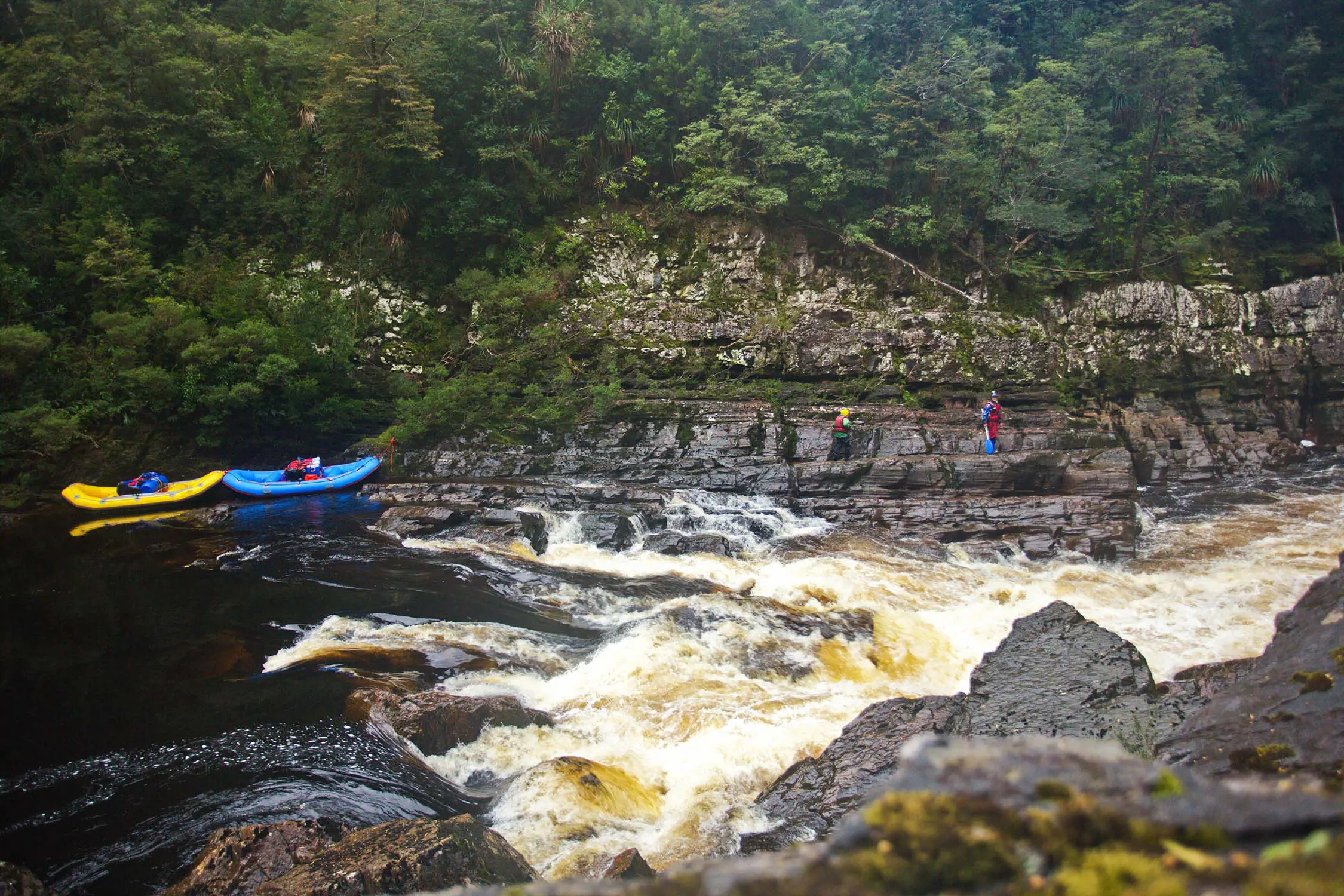 Two people and their brightly coloured canoes stand on the rocky banks of a rapidly flowing river.