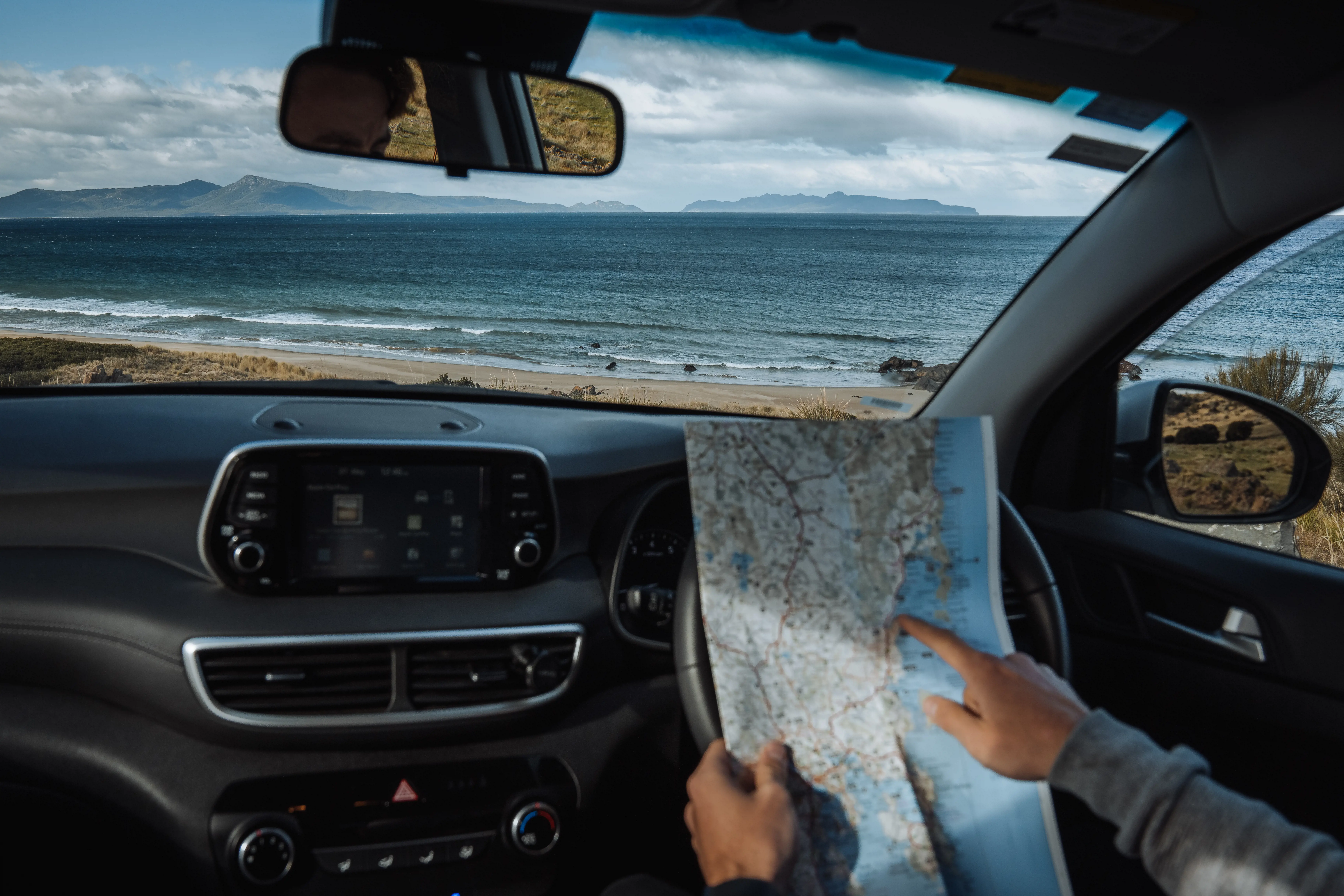 A man sits in his car, parked by the seaside, pointing at a map