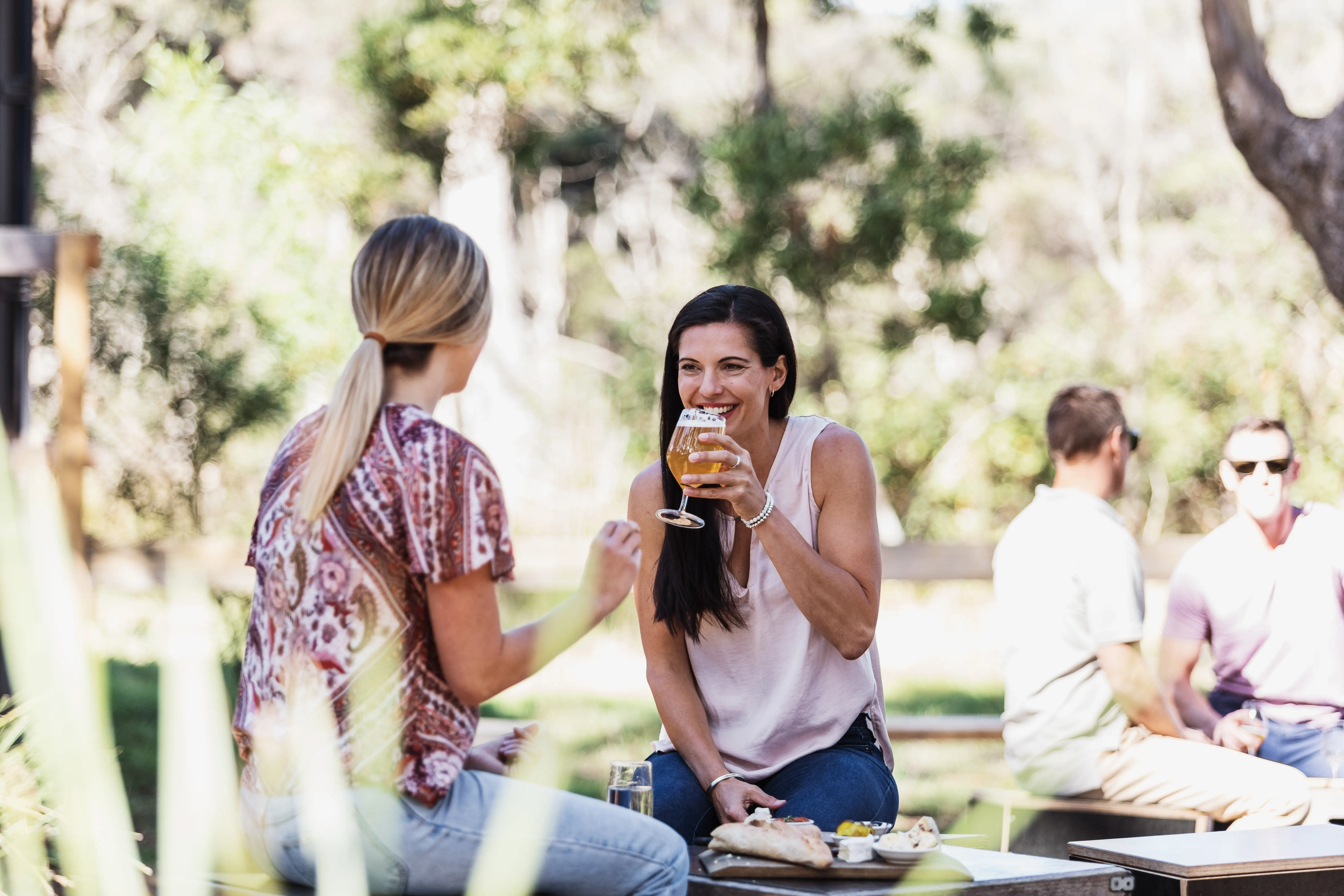 Two people sit laughing and talking on low stools under trees, drinking from beer glasses.