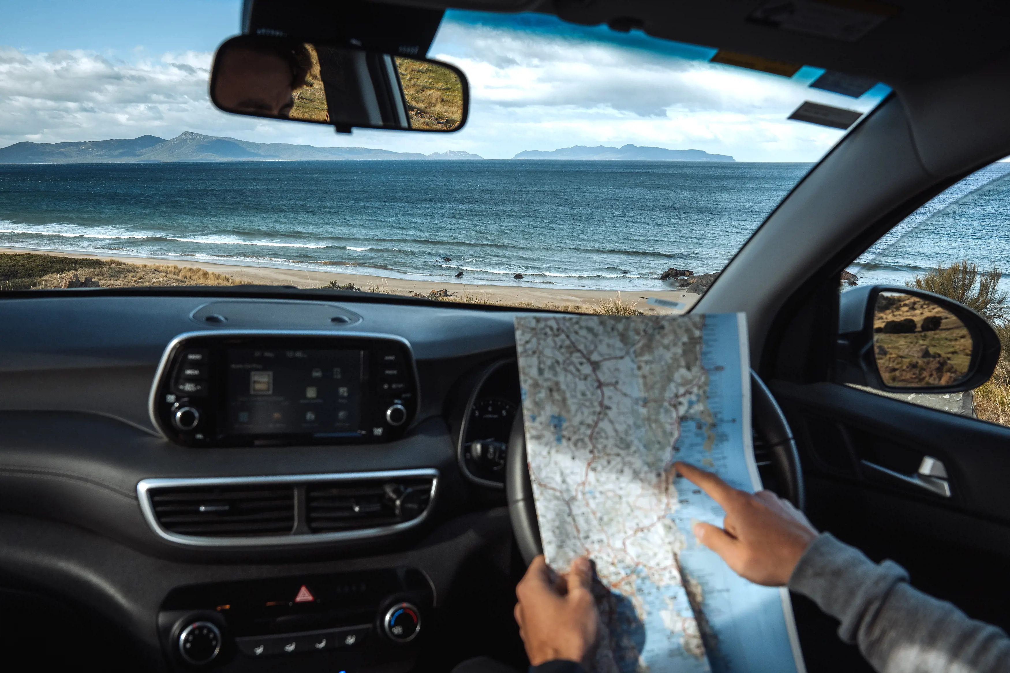 A man sits in his car, parked by the seaside, pointing at a map