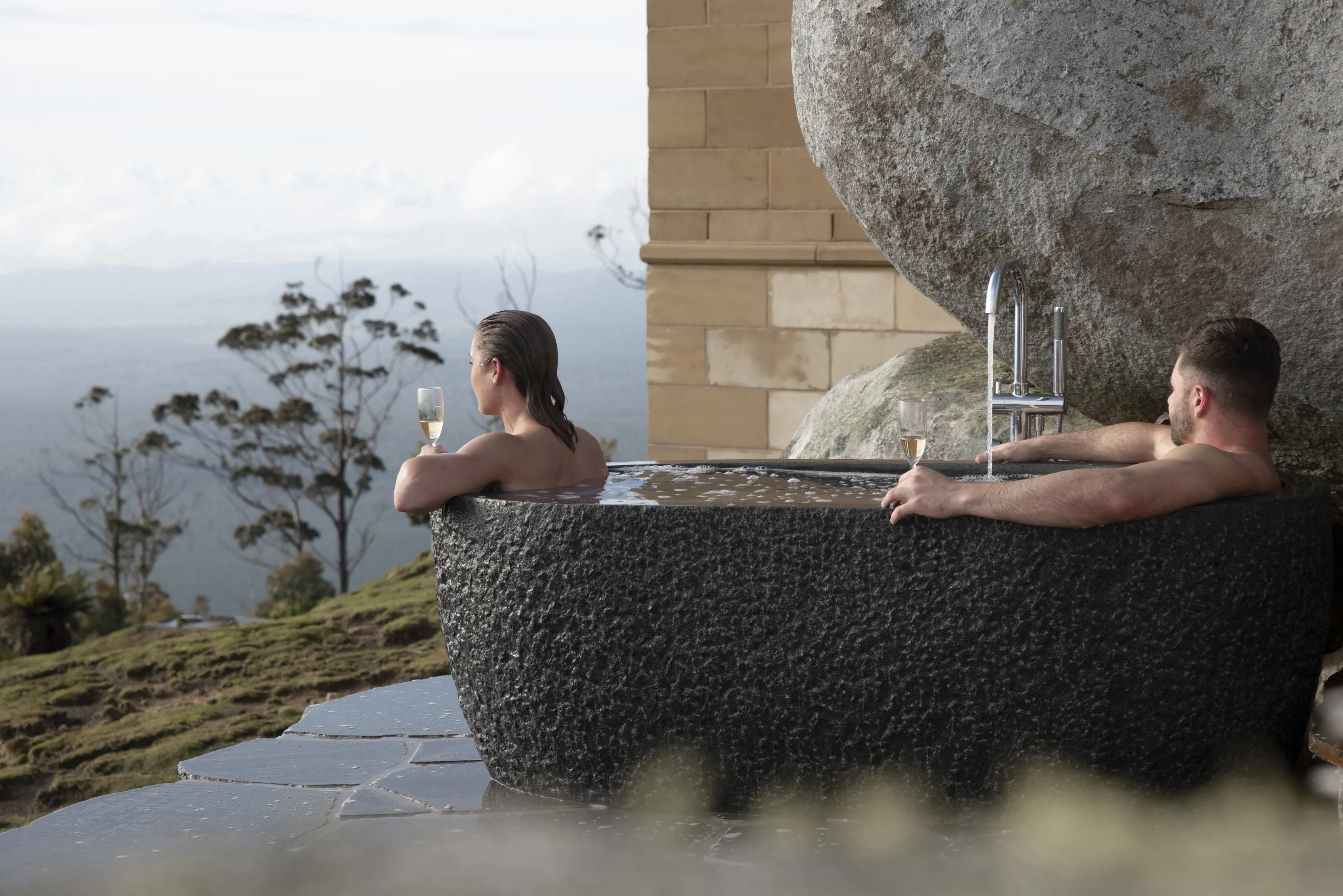 A couple relax with champagne in a hot outdoor bath at The Keep, perched upon a 650 metre rocky pinnacle overlooking Tasmania’s North east.