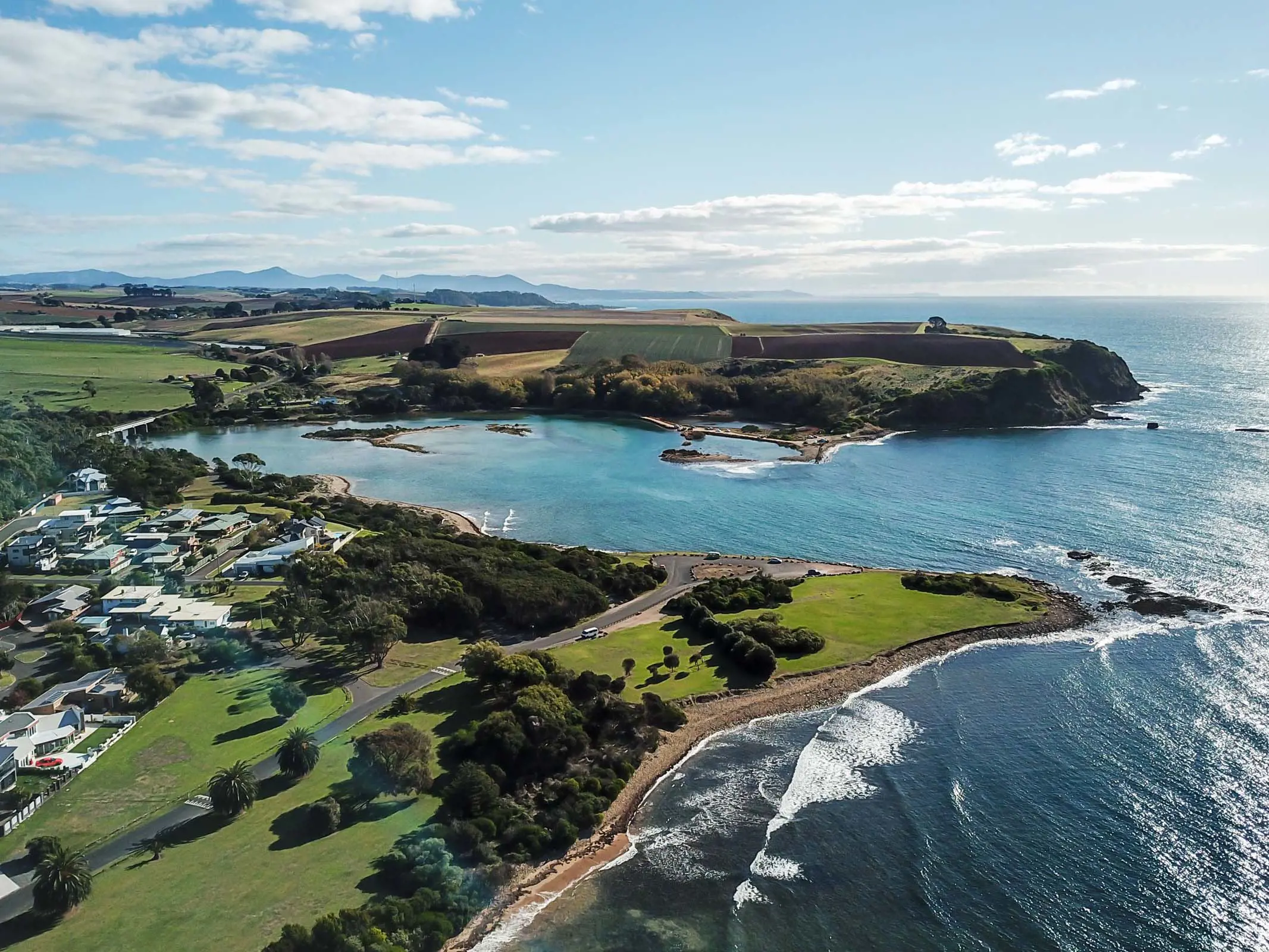 An aerial view of a coastline and beaches, surrounded by rocky bluffs. The land is richly green with trees and buildings dotted throughout.