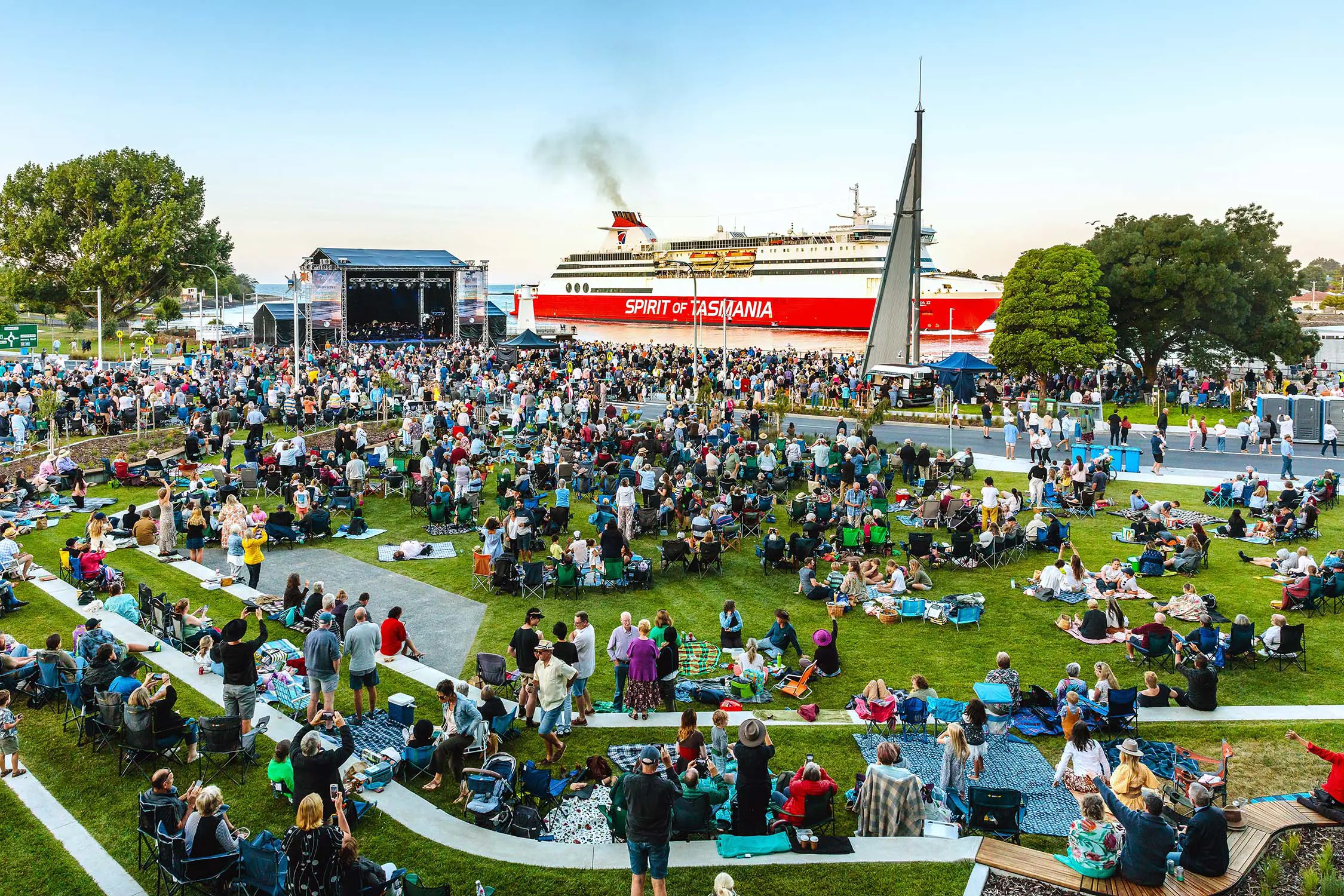 A view of a packed waterfront park grassed area, with people on camp chairs and picnic blankets. An outdoor stage is set up to one side, and the bright red Spirit of Tasmania ship sits prominently in the background.